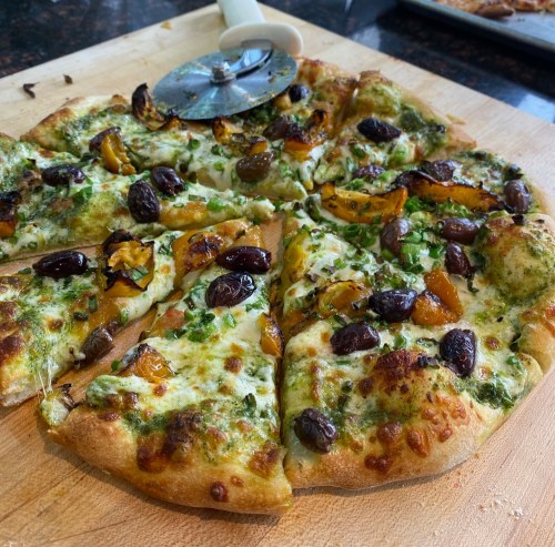 A freshly baked pizza topped with pesto, olives, and roasted vegetables, sliced and ready to serve.