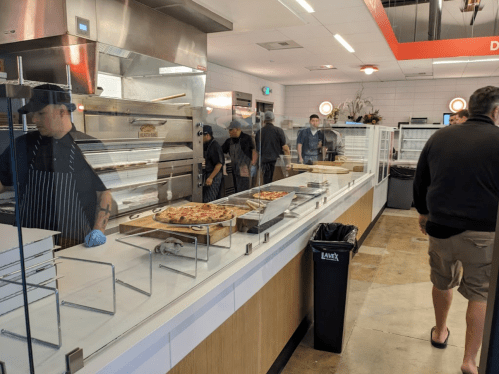 A busy pizza restaurant with chefs behind the counter and customers in line, showcasing various pizzas on display.