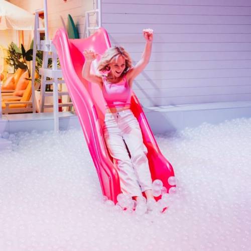 A girl in a pink top joyfully slides down a bright pink slide into a pool of white plastic balls.