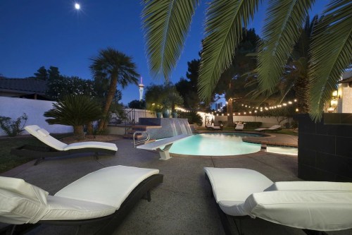 A serene backyard at night featuring a pool, lounge chairs, palm trees, and string lights illuminating the space.