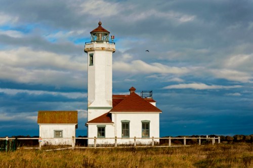 A white lighthouse with a red roof stands against a cloudy sky, surrounded by grassy land and a stone wall.