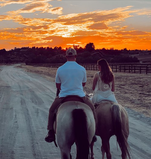 A couple rides horses down a dirt path at sunset, with vibrant orange and purple skies in the background.