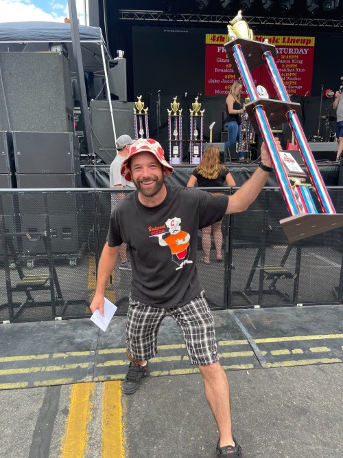 A man in a bucket hat and t-shirt holds a large trophy, smiling and posing in front of a stage with trophies in the background.