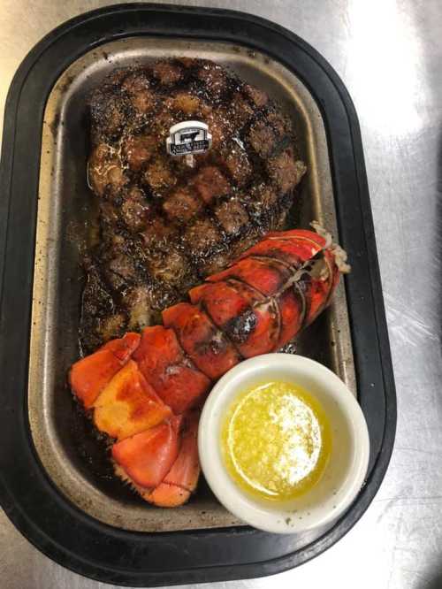 A grilled steak with a lobster tail and a small bowl of melted butter on a metal tray.