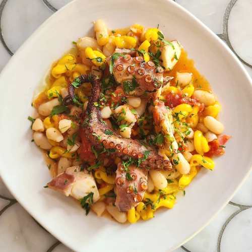 A bowl of octopus with white beans, corn, and herbs, garnished with fresh parsley on a marble surface.