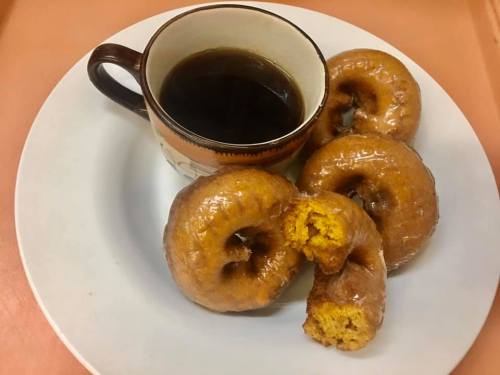 A plate with four glazed donuts and a cup of black coffee. One donut is partially bitten.