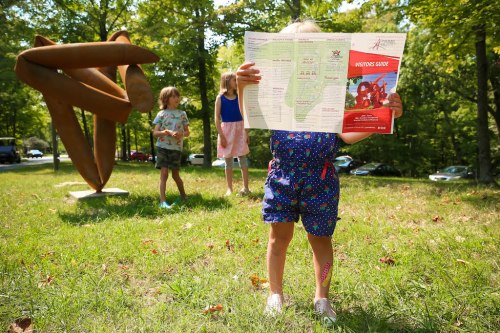 A child holds a map in a park, with two other kids and a large sculpture in the background among green trees.
