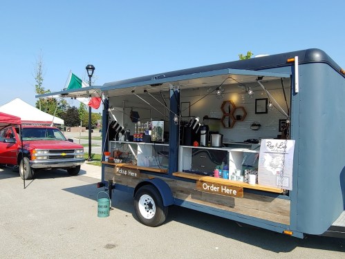 A food truck with a "Pickup Here" sign, serving drinks and snacks, parked at an outdoor event with tents nearby.