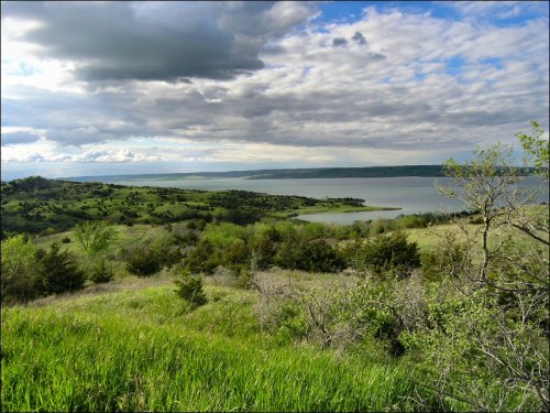 A scenic view of a lake surrounded by green hills and trees under a cloudy sky.