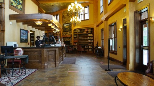 A cozy hotel lobby with wooden decor, a reception desk, bookshelves, and large windows letting in natural light.