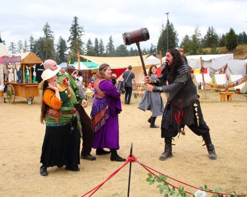 A medieval fair scene with costumed characters, including a man wielding a large hammer and women in colorful outfits.