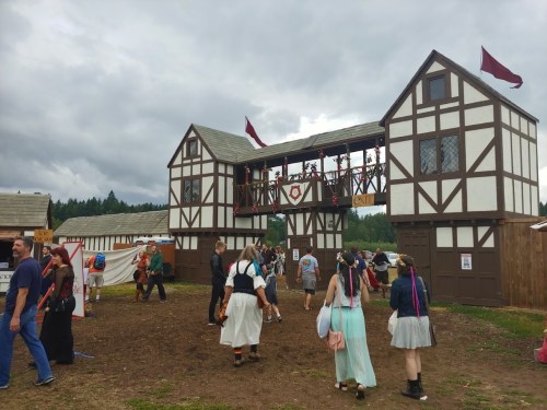 A medieval-themed festival scene with people walking past a large, wooden building adorned with flags and decorations.
