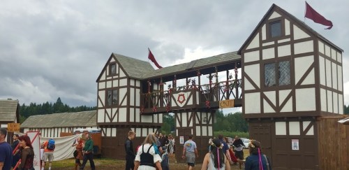 A medieval-style entrance with wooden beams and flags, bustling with people at a festival.