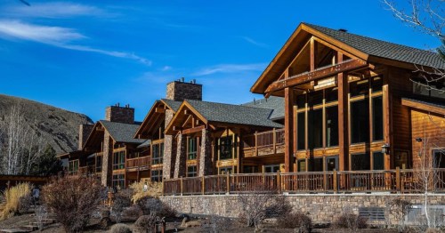 A row of rustic wooden buildings with large windows, set against a clear blue sky and mountainous backdrop.