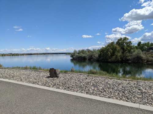 A calm river reflects the sky, bordered by greenery and a pebbled path under a blue sky with fluffy clouds.