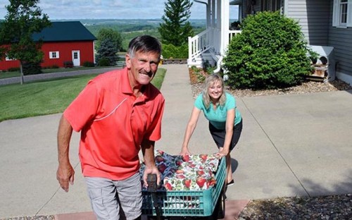 A man and woman smile while moving a cart filled with colorful blankets on a sunny day outside a house.