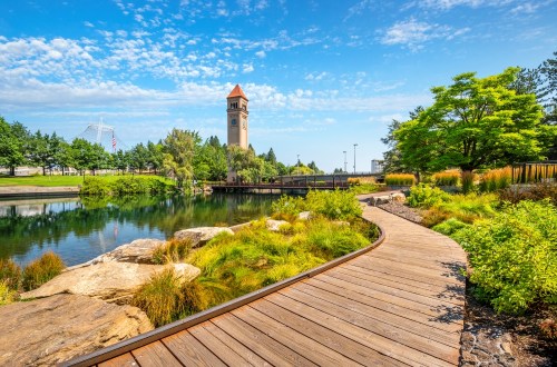 A scenic park with a wooden path, a clock tower, lush greenery, and a calm pond under a blue sky.