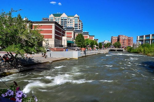 A river flows past buildings and greenery under a clear blue sky, with people enjoying the waterfront.