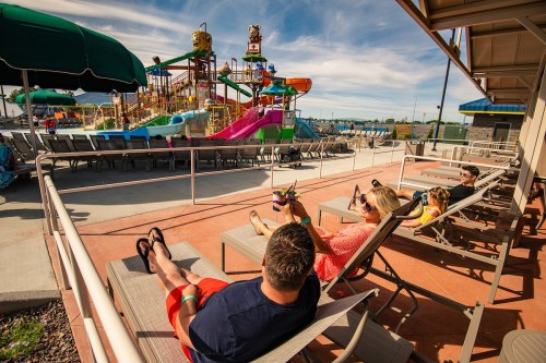 A group relaxes on lounge chairs by a water park with colorful slides and attractions in the background.