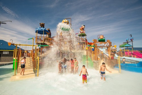Children and adults enjoy a water park splash pad with colorful slides and a large water feature on a sunny day.
