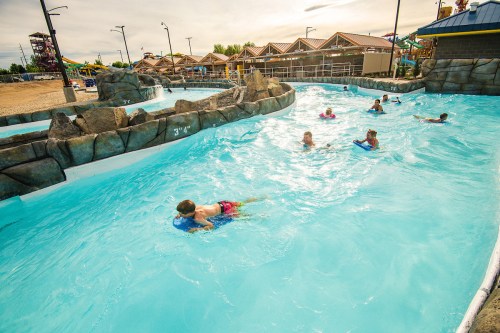 A sunny water park scene with children swimming and floating on rafts in a blue lazy river.