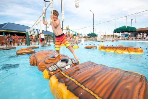 A boy balances on a floating lion in a pool, with people enjoying the water and sun in the background.