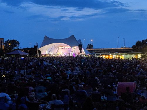 A large crowd gathers at an outdoor concert under a tent, illuminated by stage lights against a twilight sky.