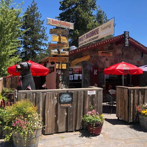 A rustic restaurant with a wooden fence, red umbrellas, and a bear statue outside, surrounded by trees and signs.