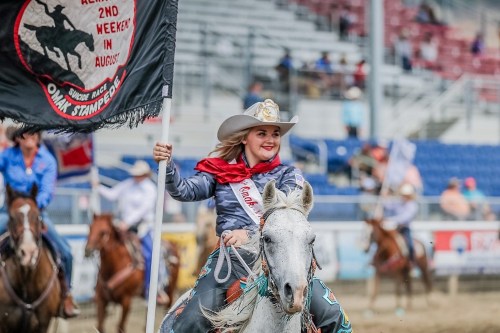 A smiling woman in a cowboy hat holds a flag while riding a white horse at a rodeo event.