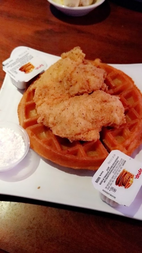 A plate featuring a waffle topped with two pieces of fried chicken and two packets of syrup.
