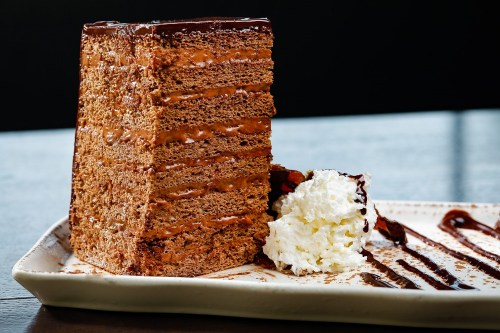 A tall slice of chocolate cake with layers, accompanied by whipped cream and chocolate drizzle on a plate.