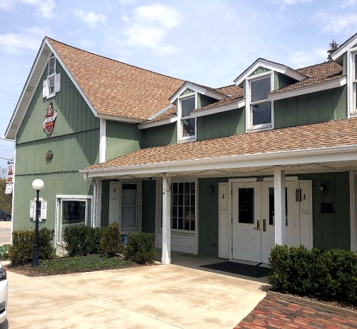 A green building with a brown roof, featuring a sign and large windows, surrounded by shrubs and a paved entrance.