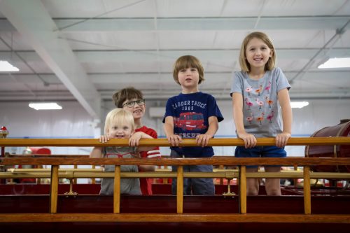 Four children smile and lean on a wooden railing inside a bright, spacious building.