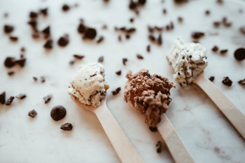 Three scoops of ice cream on wooden spoons, surrounded by chocolate chips on a marble surface.