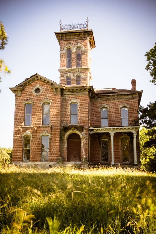 An abandoned brick mansion with a tall tower, surrounded by overgrown grass and trees under a clear sky.