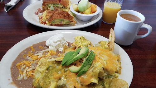 A plate of nachos topped with eggs, cheese, and avocado, alongside a fruit cup and a croissant sandwich. Coffee in a cup.