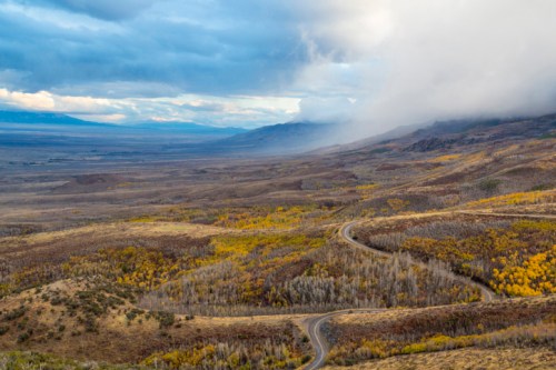 A winding road through a valley with autumn foliage and mountains under a cloudy sky.