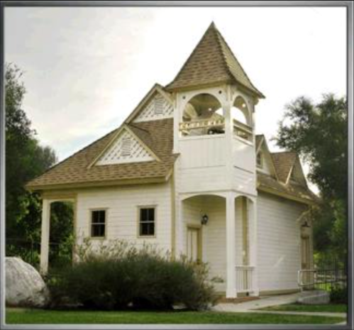 A charming white house with a unique tower and a sloped roof, surrounded by greenery and a large rock.