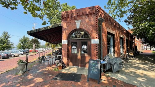 A brick building with large windows and outdoor seating, surrounded by trees and a view of the water.
