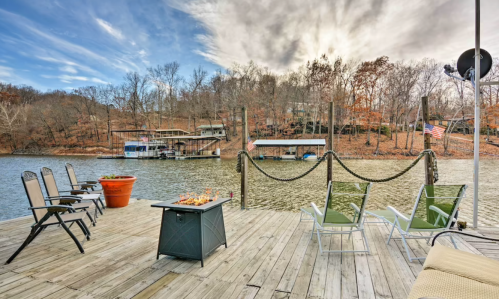 A serene lakeside view with a wooden dock, chairs, a fire pit, and autumn foliage in the background.