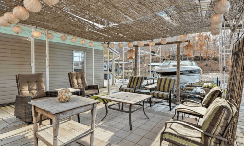 Cozy outdoor patio with seating, a table, and a boat in the background, shaded by a woven canopy and string lights.