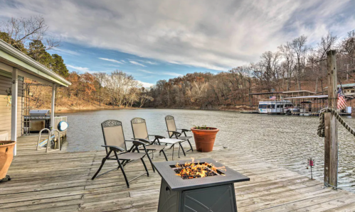A serene lakeside scene with chairs, a fire pit, and trees reflecting autumn colors under a cloudy sky.