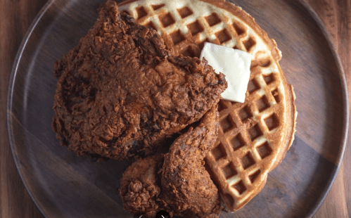 A wooden plate with crispy fried chicken and a golden waffle topped with a pat of butter.
