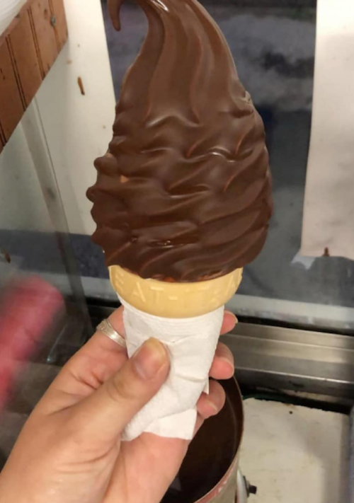 A hand holding a chocolate-dipped soft serve ice cream cone with a paper napkin wrapped around the cone.