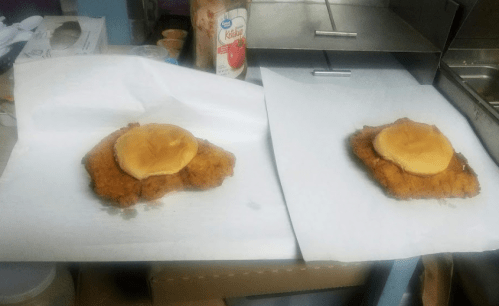 Two fried chicken patties on paper, each topped with a slice of cheese, on a kitchen counter.
