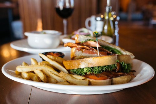 A close-up of a sandwich with bacon, lettuce, and tomato, served with fries and a glass of red wine in the background.