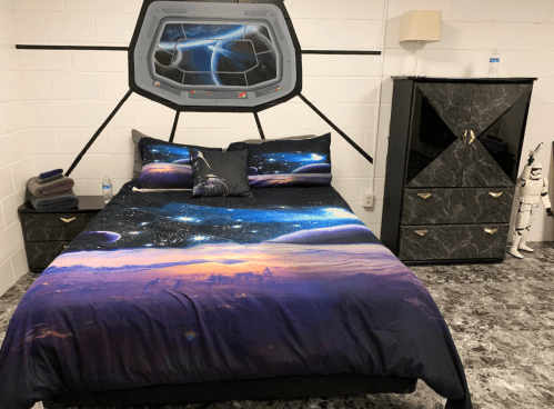 A space-themed bedroom featuring a galaxy print bedspread, decorative pillows, and a modern dresser against a white wall.