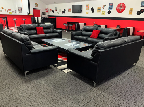 A retro-style lounge with black leather sofas, red cushions, and a glass coffee table on a patterned rug.
