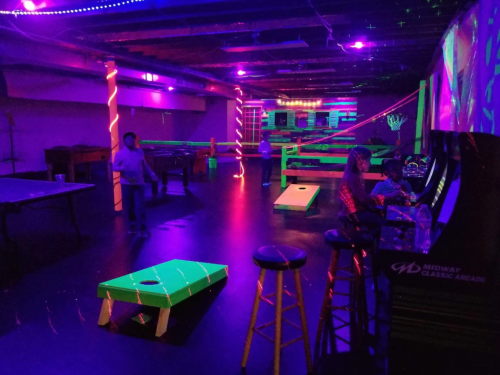 A dimly lit arcade with neon lights, featuring pool tables, cornhole, and arcade games. People are enjoying the space.
