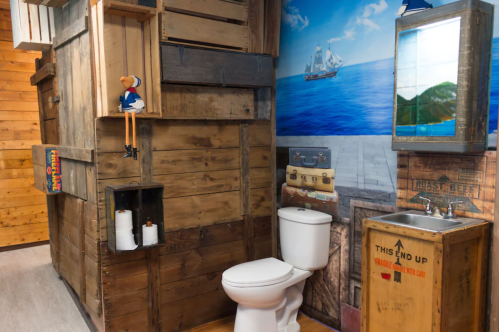 A rustic bathroom with wooden walls, a toilet, and nautical-themed decor, including a ship mural and a wooden shelf.